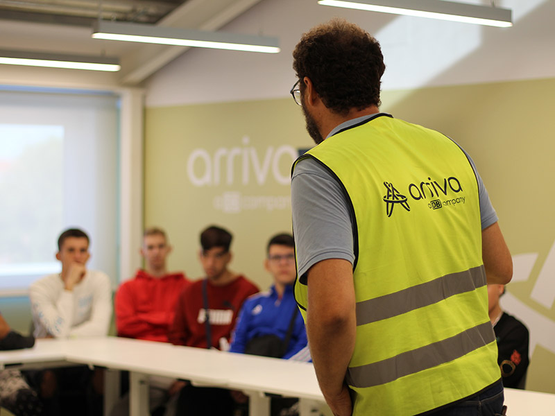 Un hombre, con un chaleco reflectante con el logo de Arriva, habla con un grupo de jóvenes sentados en una mesa.