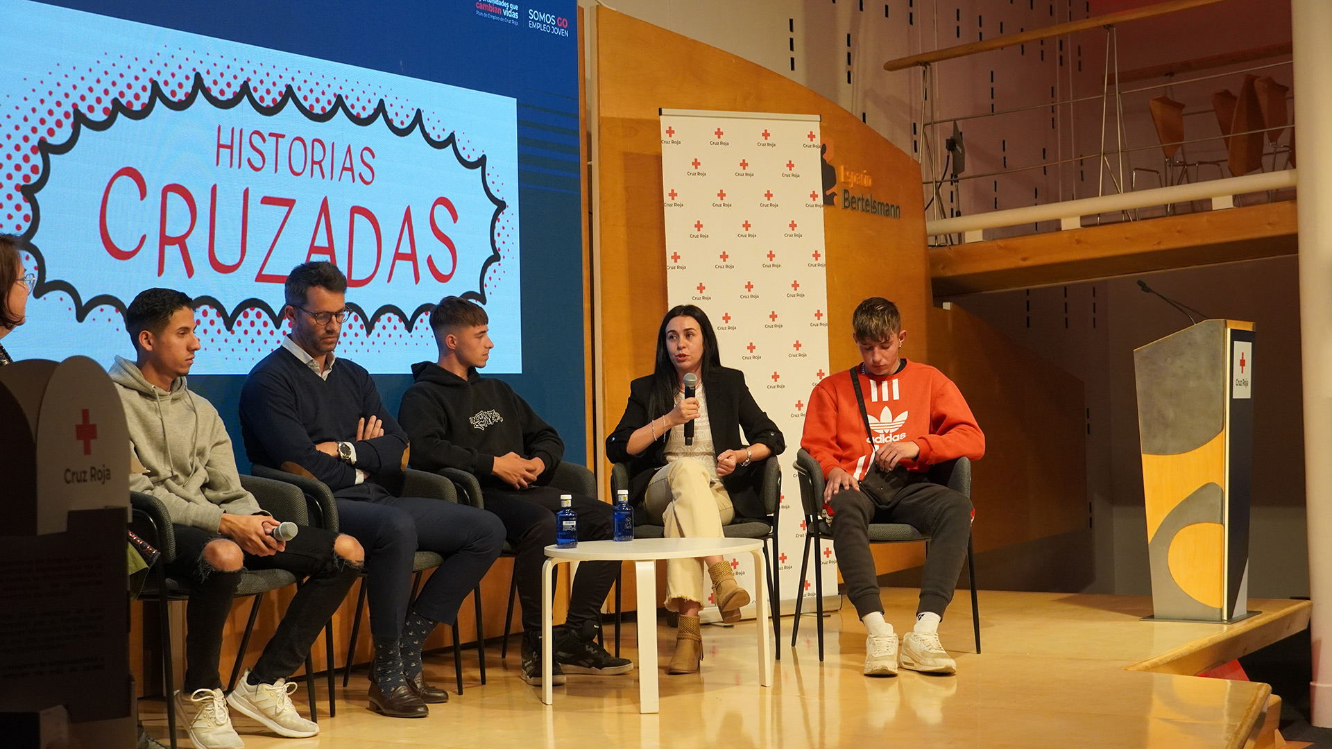 Una mujer con un micrófono de mano participa en una charla rodeada de jóvenes, frente a una pantalla que indica "Historias cruzadas"