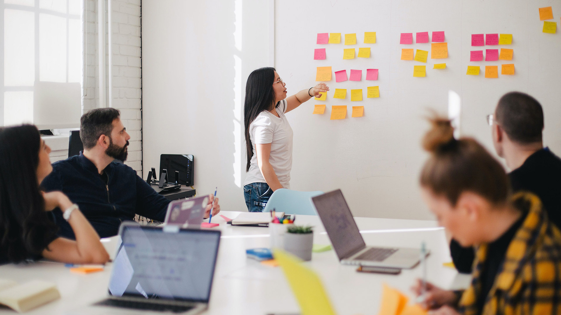 Varias personas sentadas a una mesa, con ordenadores portátiles, escuchan a una persona de pie junto a una pizarra llena de post its de colores.