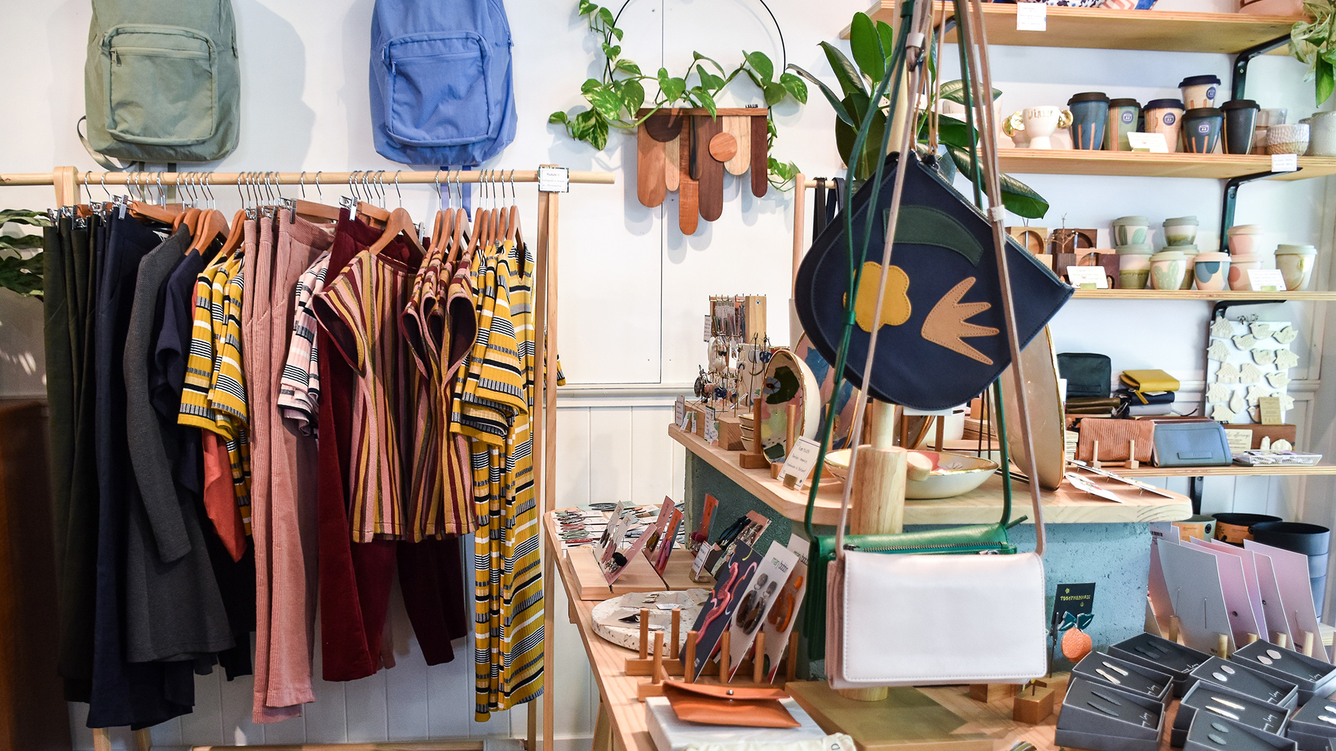 Interior de una tienda donde se ven dos mochilas colgadas de la pared, camisetas y pantalones en un perchero, y bolsos y carteras sobre una mesa.