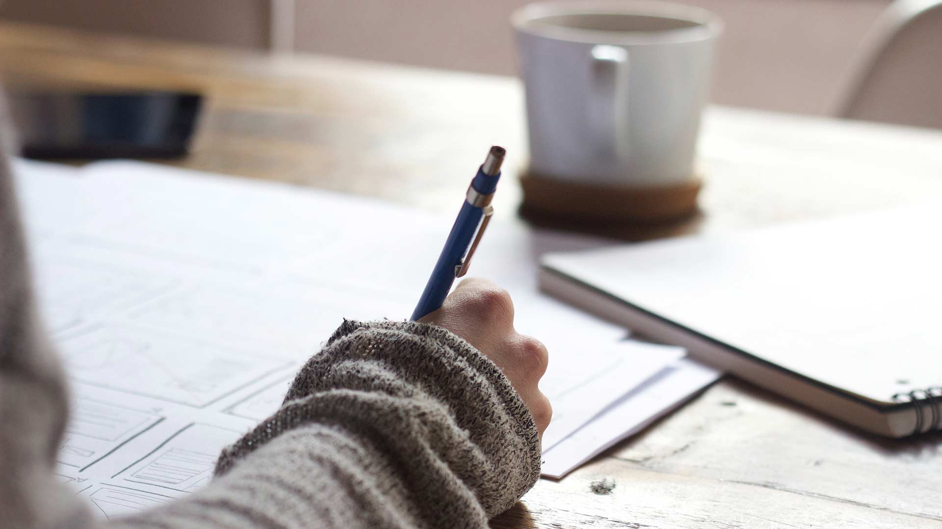 Una mesa con un cuaderno, una taza y apuntes que una persona está estudiando con un bolígrafo en la mano.
