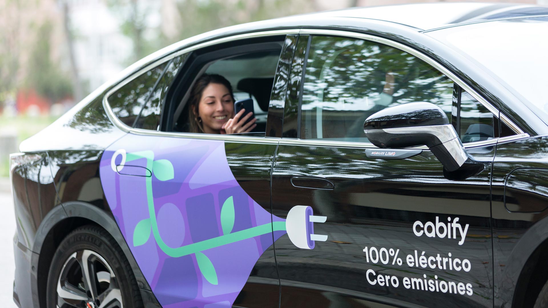 Un coche negro vinilado con el texto: Cabify 100% eléctrico Cero emisiones. Dentro, una chica mira el móvil desde el asiento trasero