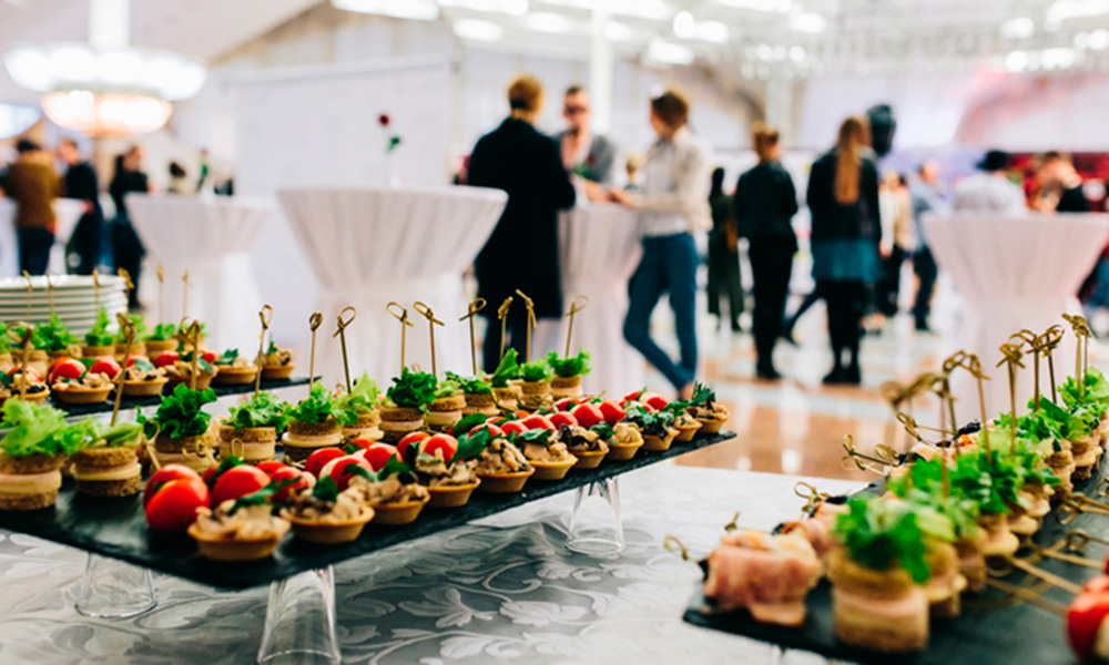 Mesa de aperitivos en un catering. Al fondo, varias personas se agrupan en torno a mesas altas.