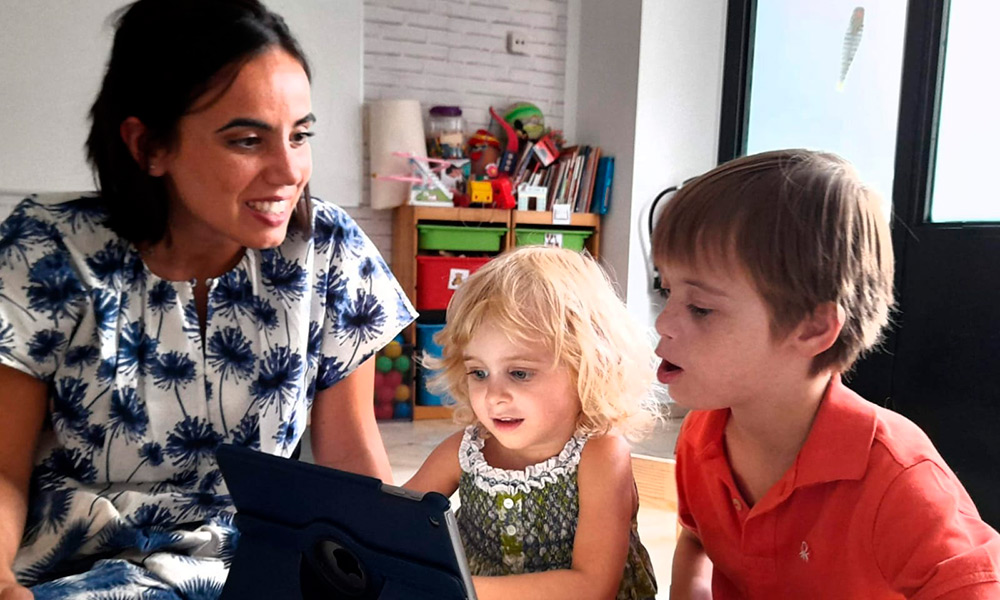 Una mujer interactúa con dos niños estudiando frente a una pantalla de ordenador.